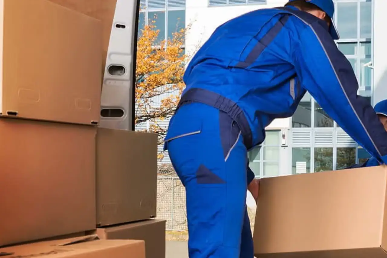 Two delivery workers in blue uniforms are loading cardboard boxes into a van outside a building. Trees are visible in the background Two delivery workers in blue uniforms are loading cardboard boxes into a van outside a building. Trees are visible in the background
