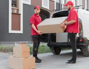 Two delivery workers in red shirts are transferring a large cardboard box from a van to the ground, with stacked boxes nearby Two delivery workers in red shirts are transferring a large cardboard box from a van to the ground, with stacked boxes nearby